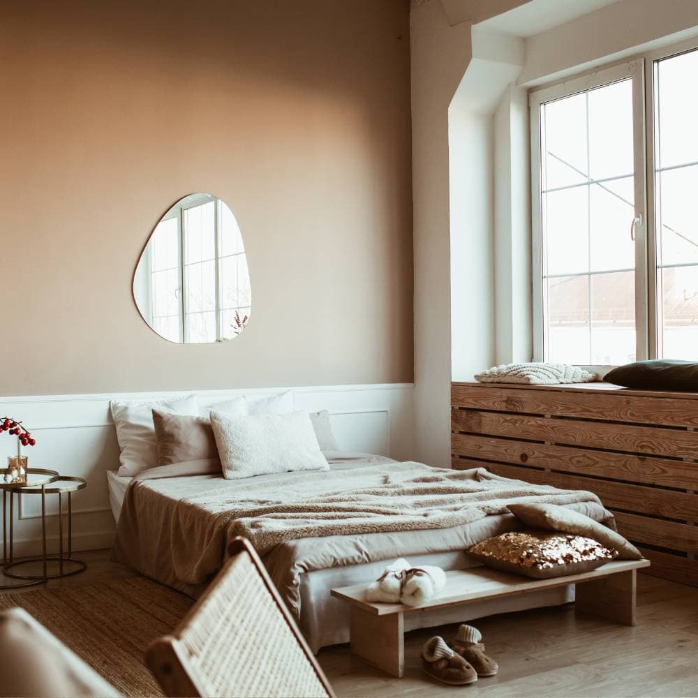 Cozy bedroom with a low bed, beige bedding, abstract wall mirror, and large window. A rustic wooden bench and slippers are in the foreground.