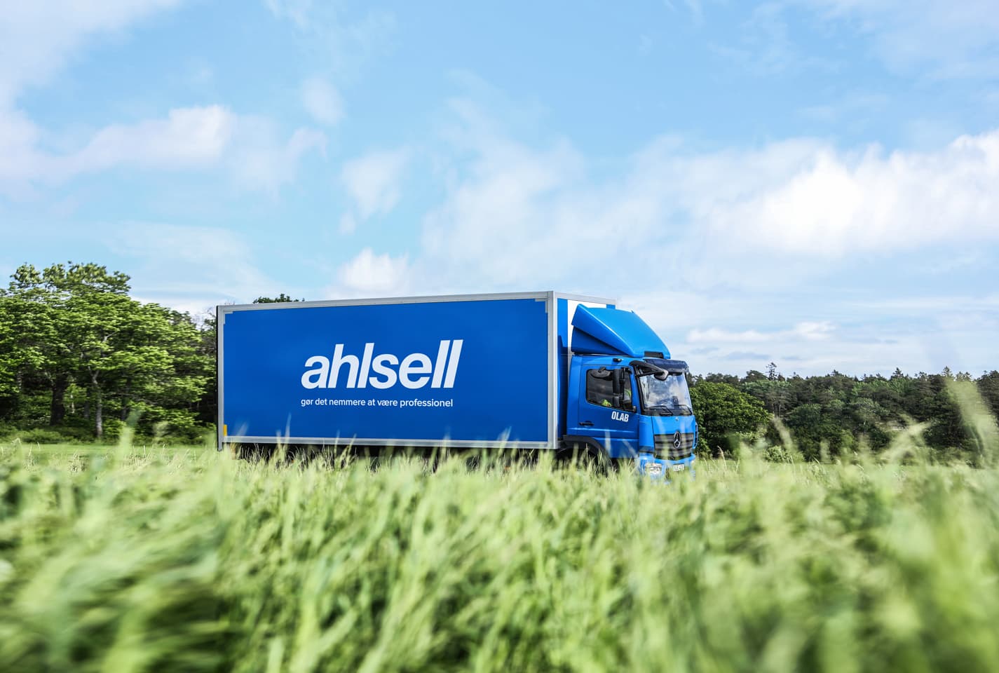 Blue delivery truck with "Ahlsell" logo drives through a lush green field under a partly cloudy sky.
